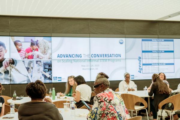 Small group of people talking at tables in front of large screen that says Advancing the converstation on scaling national informal STEM programs