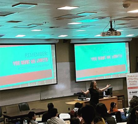 Woman speaking in front of a group of students. Screens behind her read "Remember: You have no limits"