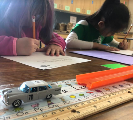 Two girls writing on pieces of paper. In the foreground, a matchbox car and track.