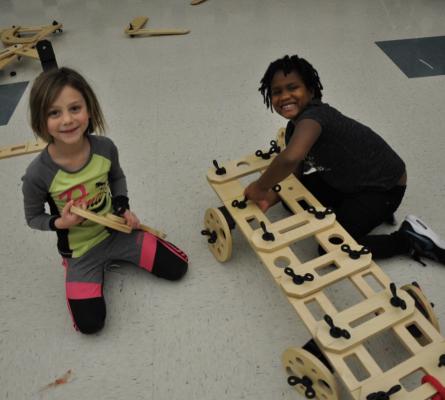 Two girls building a large car with wooden pieces from a rig a ma jig set