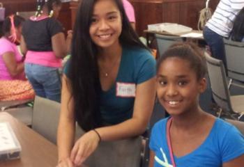 Photo of two young women, both wearing blue shirts, at a table with robotics parts on it