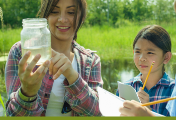 Woman with young girl looking at jar of water in front of pond. Girl is holding a notebook and pencil