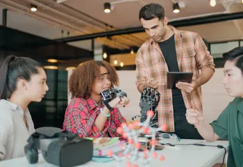 Group of young adults sitting a table with various tech products on it.