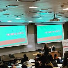 Woman speaking in front of a group of students. Screens behind her read "Remember: You have no limits"