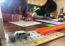 Two girls writing on pieces of paper. In the foreground, a matchbox car and track.