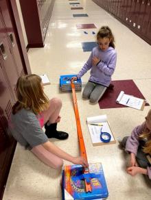 Three girls sitting on the floor next to an orange matchbox car track