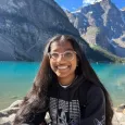 Maithili Kumar - Young woman with long dark hair and glasses sitting in front of a lake with mountains behind
