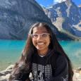 Maithili Kumar - Young woman with long dark hair and glasses sitting in front of a lake with mountains behind