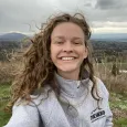 A selfie of Emma Hagen, who has white skin, grey eyes, and curly light-brown hair. She is smiling at the camera. Her hair is blowing in the wind and behind her is a scene of mountains, an open field, and evergreen and deciduous trees. 