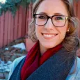 Joanne Esch - woman with brown hair and glasses wearing a sweater and scarf standing in front of a red fence