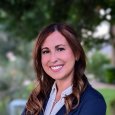 Alyson Freeman - Woman with brown hair wearing blue blazer and white shirt standing in front of trees and shrubs