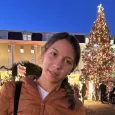 Teen girl wearing a brown jacket standing in front of a large decorated Christmas tree