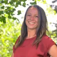 Heather Jameson headshot - photo of woman with brown hair wearing a red top standing in front of trees