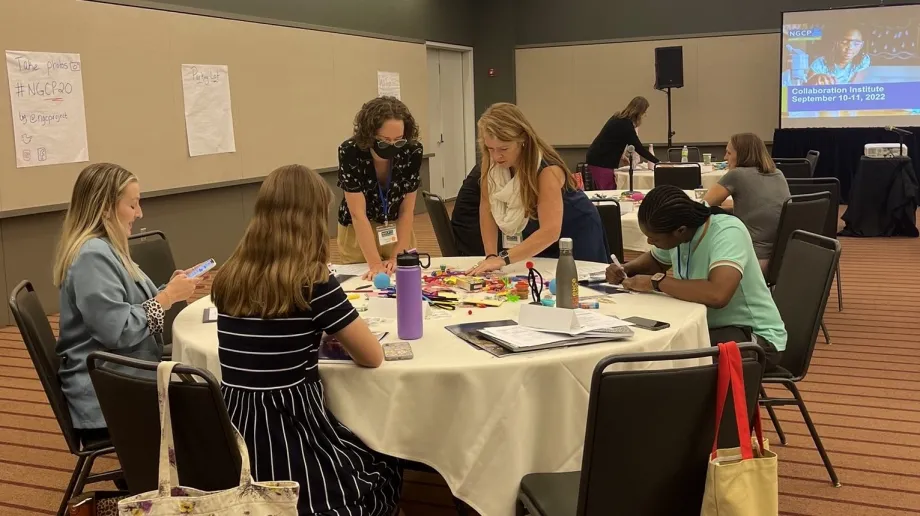 A photo showing a group of five people surrounding a table and appearing to work together on a project. There is a collection of various supplies covering the table